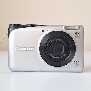 May include: A silver Canon digital camera with a black lens and a 4x optical zoom. The camera has a strap attached to it.