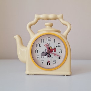 May include: A yellow ceramic teapot shaped clock with a black and white illustration of a train and a red castle on the clock face. The clock face has black numbers and a yellow rim.