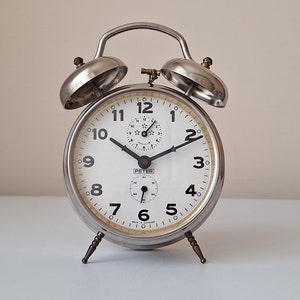 Könnte beinhalten: Ein silberner Wecker mit weißem Zifferblatt und schwarzen Zahlen. Die Uhr hat einen kleinen Sekundenzeiger und einen größeren Minuten- und Stundenzeiger. Die Uhr ist auf 7:55 eingestellt.