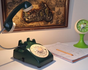 May include: A green rotary phone with a flexible arm and a white receiver. The phone is on a white desk with a notebook and pencil, and a green clock with a white face.