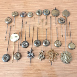 May include: A collection of antique clock pendulums in various shapes and sizes. The pendulums are made of metal and have a variety of decorative elements, including sunbursts, figures, and ornate designs. The pendulums are arranged on a light brown wooden surface.