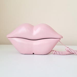 May include: A light pink novelty telephone shaped like a pair of lips. The phone has a matching pink coiled cord. The phone is a retro design and sits on a white surface.