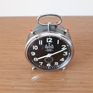 Puede incluir: Un reloj despertador plateado con una esfera negra y números blancos. El reloj tiene una manija cromada y un interruptor de alarma rojo y verde. La esfera del reloj dice "Wehle Three in One" y "Jewelled".