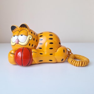 Op de afbeelding: Een vintage Garfield-telefoon in heldergeel met zwarte stippen. De telefoon houdt een rode basketbal vast en heeft een opgerold gele snoer. Een verzamelobject.