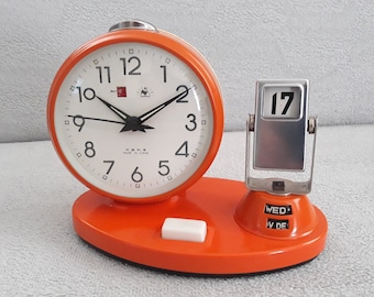 Könnte beinhalten: Orangefarbener Wecker mit weißem Zifferblatt und schwarzen Zeigern. Die Uhr verfügt über eine Kalenderfunktion mit einem silbernen Klappdisplay, das das Datum "17" und den Wochentag "WED" anzeigt.