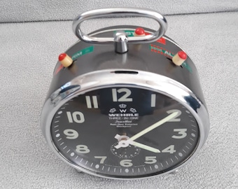 May include: A silver alarm clock with a black face and white numbers. The clock has a silver handle on the top and a red and green button on each side. The clock face reads "WEHRLE THREE-IN-ONE Jewelled Rust & Dust Protected Shockproof".