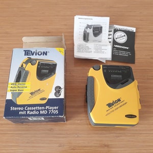 Könnte beinhalten: Gelber und grauer Kassettenspieler mit Radio. Der Spieler trägt den Text "TEVION" und "WATER RESISTANT". Die Schachtel für den Spieler trägt den Text "TEVION" und "Stereo Cassetten-Player mit Radio MD 7705".