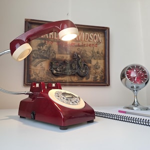 May include: A vintage-style desk lamp shaped like a rotary phone, in a deep red color with a cream-colored dial and handset. The lamp has a flexible, silver-colored gooseneck and a small lightbulb. A framed Harley-Davidson poster is in the background.