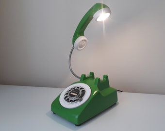 May include: A green rotary phone with a white dial and a white phone receiver that has a light bulb attached to it. The phone is on a white surface.
