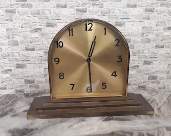 May include: A gold-toned analogue clock with black Roman numeral hour markers and black hands. The clock is set to 12:00 and is sitting on a wooden base.