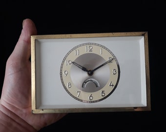 May include: A white clock with black hands and gold trim. The clock face has black Roman numerals and a silver dial with the word "Noveda" printed on it.