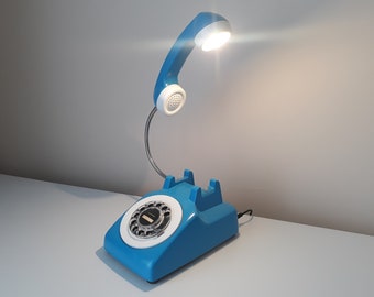 May include: A blue rotary phone shaped desk lamp with a white dial and a white light bulb. The lamp is on a white surface.