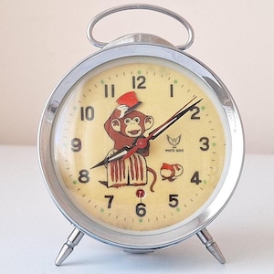May include: A vintage silver alarm clock with a yellow face and a cartoon monkey wearing a red hat. The clock has black numbers and hands, and the words "White Bird" are printed on the face.