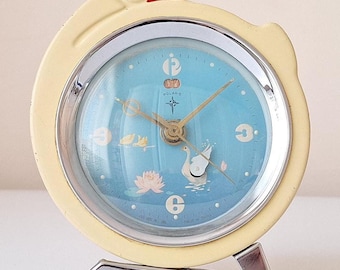 Könnte beinhalten: Ein gelber, schwanenförmiger Wecker mit blauem Zifferblatt und silbernen Verzierungen. Die Uhr hat ein weißes Schwanen- und Seerosenmotiv auf dem Zifferblatt.