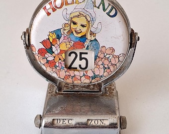 Pode incluir: Um calendário de mesa prateado com um rosto redondo branco que apresenta uma menina com um vestido azul e branco segurando flores. O rosto diz "HOLLAND" e mostra a data "25". A base do calendário tem as palavras "DEC" e "ZON" nela.