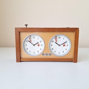 May include: A vintage wooden chess clock with two circular clock faces. Each clock face has black numerals, black hands, and a red star marker. The clock is labeled "GARDE" and has a wooden frame.