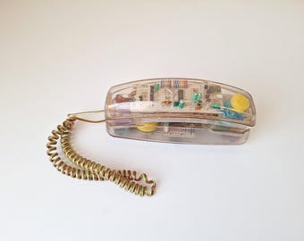 Może przedstawiać: Przezroczysty telefon w stylu vintage z widocznymi elementami wewnętrznymi. Telefon ma przezroczystą plastikową obudowę, żółtą tarczę i spiralny przewód w odcieniach zieleni i złota. Projekt prezentuje wewnętrzne obwody.