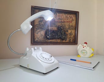 May include: A white telephone-shaped desk lamp with a flexible silver neck and a bright light in the handset. The lamp sits on a white surface, with a framed Harley-Davidson print in the background.