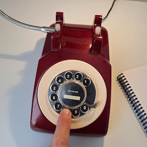 May include: A vintage burgundy rotary dial telephone with a cream-colored dial and black numbers. A finger presses the number seven. A spiral notebook sits to the right. A flexible metal tube extends from the phone.