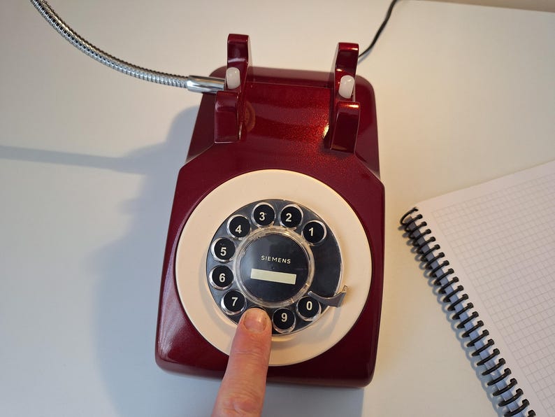 May include: A vintage burgundy rotary dial telephone with a cream-colored dial and black numbers. A finger presses the number seven. A spiral notebook sits to the right. A flexible metal tube extends from the phone.