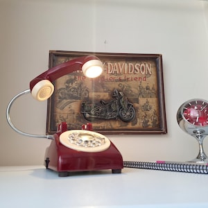 May include: A vintage burgundy telephone lamp with a cream-colored dial and handset. The lamp is next to a framed Harley-Davidson sign and a chrome clock. A spiral notebook is in the foreground.