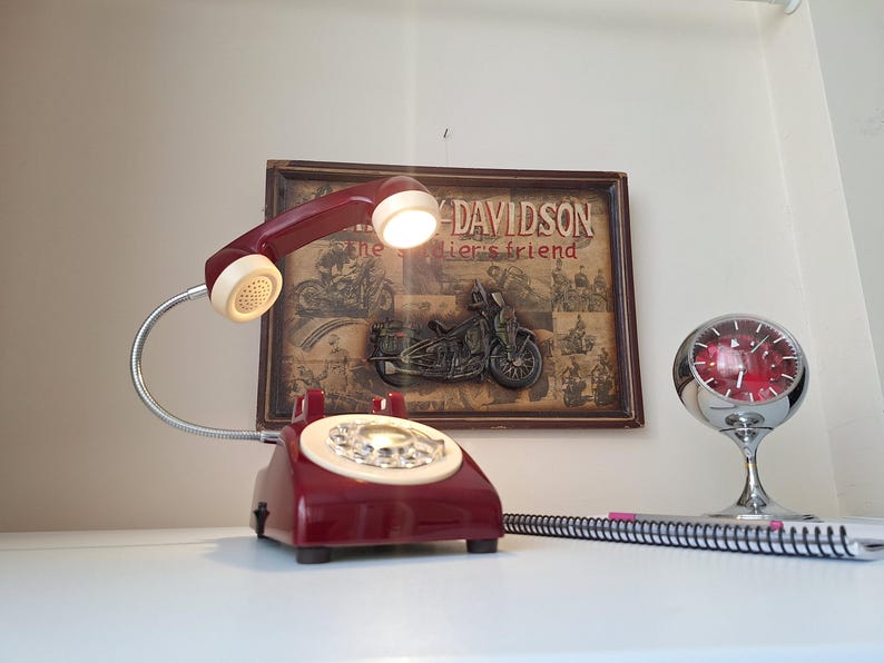 May include: A vintage burgundy telephone lamp with a cream-colored dial and handset. The lamp is next to a framed Harley-Davidson sign and a chrome clock. A spiral notebook is in the foreground.