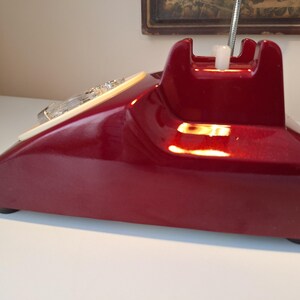 May include: A vintage, burgundy-colored telephone with a glossy finish. The rotary dial is cream-colored, and the handset cradle is a darker shade of burgundy. The phone has a classic, retro design, with a chrome-colored flexible cord.