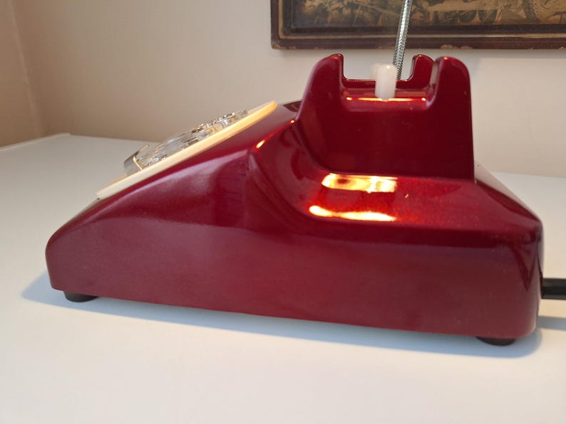 May include: A vintage, burgundy-colored telephone with a glossy finish. The rotary dial is cream-colored, and the handset cradle is a darker shade of burgundy. The phone has a classic, retro design, with a chrome-colored flexible cord.