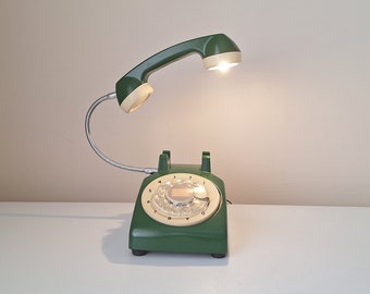 May include: A green rotary phone with a beige dial and a flexible green lamp attached to the handset. The lamp is turned on and illuminating the room.