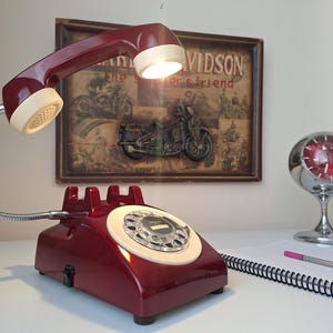 May include: A burgundy retro telephone lamp with a flexible chrome neck and a cream-colored receiver. The lamp is on a white surface, with a clock and a notepad in the background. A vintage Harley Davidson poster is behind the lamp.