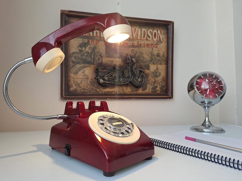 May include: A burgundy retro telephone lamp with a flexible chrome neck and a cream-colored receiver. The lamp is on a white surface, with a clock and a notepad in the background. A vintage Harley Davidson poster is behind the lamp.