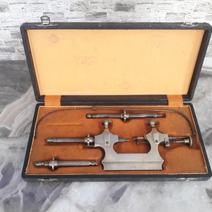 May include: A vintage metal lathe in a black case with a brown interior. The lathe has three metal rods and a metal base. The case has a metal latch.