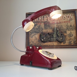 May include: A burgundy desk lamp designed as a vintage telephone. The lamp features a flexible chrome neck and a cream-colored receiver. The base has a rotary dial and a power switch. A Harley-Davidson poster is in the background.