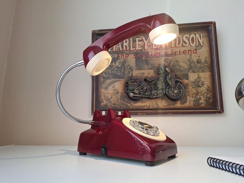 May include: A burgundy desk lamp designed as a vintage telephone. The lamp features a flexible chrome neck and a cream-colored receiver. The base has a rotary dial and a power switch. A Harley-Davidson poster is in the background.