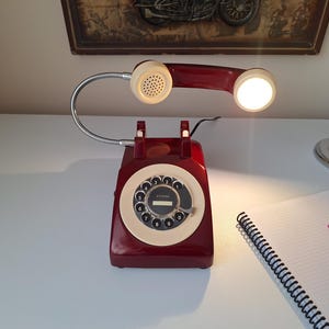 May include: A vintage-style desk lamp shaped like a rotary phone. The lamp is a deep red with a cream-colored dial and handset. The flexible neck holds a light, and the lamp sits on a white surface. A notepad and pen are nearby.