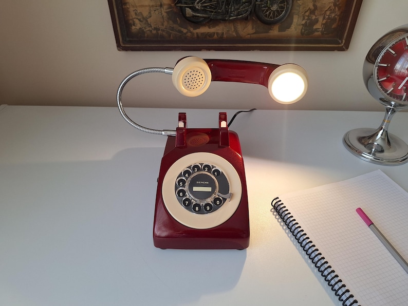 May include: A vintage-style desk lamp shaped like a rotary phone. The lamp is a deep red with a cream-colored dial and handset. The flexible neck holds a light, and the lamp sits on a white surface. A notepad and pen are nearby.