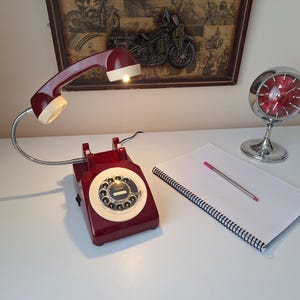 May include: A vintage-style desk lamp shaped like a rotary phone, in burgundy and cream. A chrome clock and a notepad with a pen are also on the white surface. A framed motorcycle art piece hangs in the background.