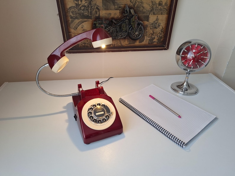 May include: A vintage-style desk lamp shaped like a rotary phone, in burgundy and cream. A chrome clock and a notepad with a pen are also on the white surface. A framed motorcycle art piece hangs in the background.