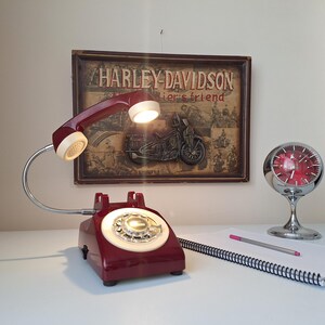 May include: A vintage-style desk lamp shaped like a rotary phone, in a deep red color, with a cream-colored handset and dial. The lamp is paired with a chrome clock and a notepad with a pencil. A Harley-Davidson sign is in the background.