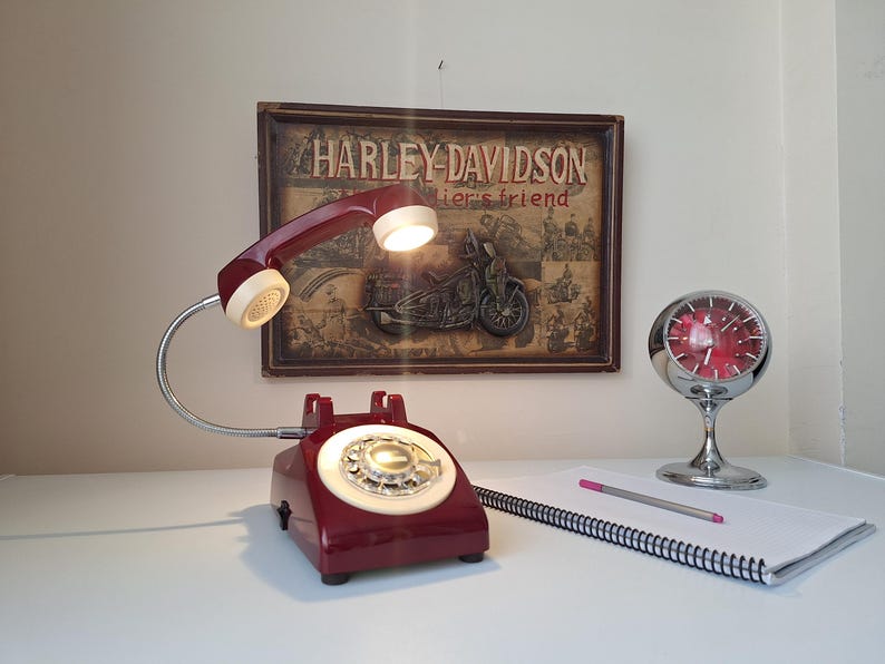 May include: A vintage-style desk lamp shaped like a rotary phone, in a deep red color, with a cream-colored handset and dial. The lamp is paired with a chrome clock and a notepad with a pencil. A Harley-Davidson sign is in the background.