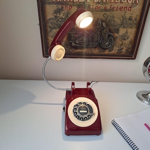 May include: A burgundy rotary phone lamp with a cream-colored dial and handset. The lamp has a flexible metal neck and a bright light. A vintage Harley-Davidson poster is in the background.