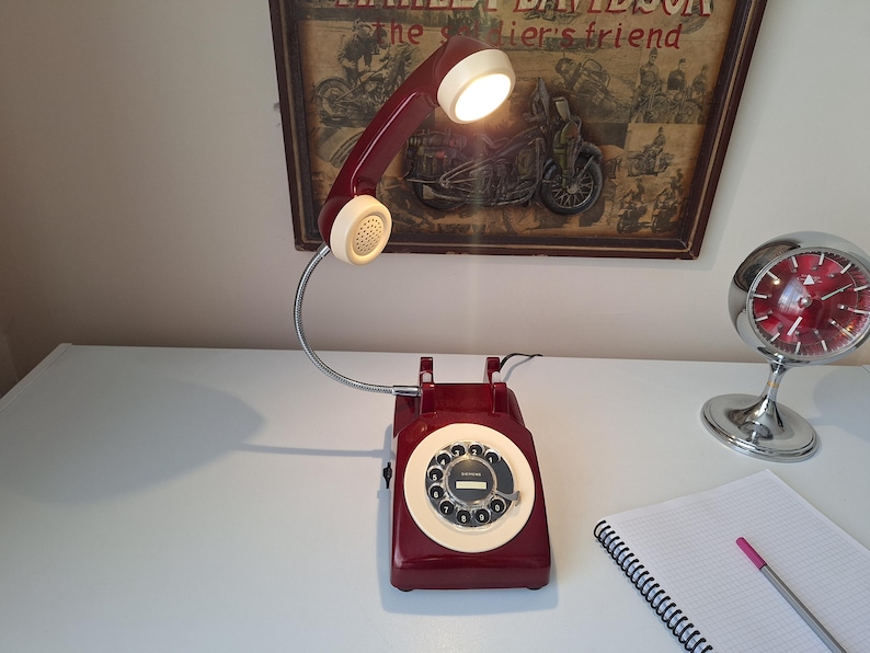 May include: A burgundy rotary phone lamp with a cream-colored dial and handset. The lamp has a flexible metal neck and a bright light. A vintage Harley-Davidson poster is in the background.