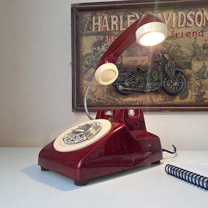 May include: A vintage burgundy telephone lamp with a cream-colored dial and handset. The lamp has a built-in light and is placed on a white surface. A framed Harley Davidson print hangs in the background.