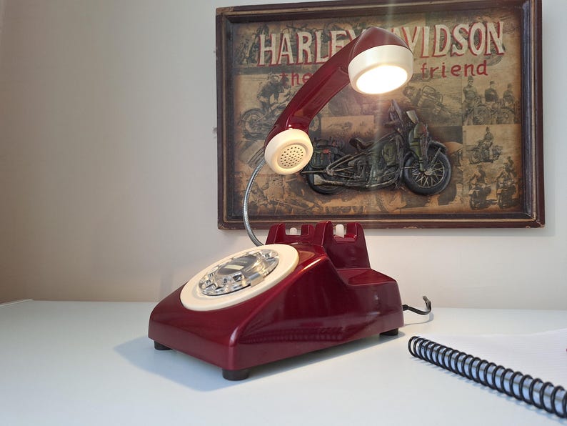 May include: A vintage burgundy telephone lamp with a cream-colored dial and handset. The lamp has a built-in light and is placed on a white surface. A framed Harley Davidson print hangs in the background.