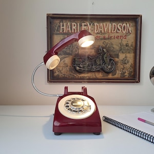 May include: A burgundy rotary phone lamp with a cream-colored dial and handset. The lamp has a flexible silver gooseneck and a small light in the handset. A Harley-Davidson sign is in the background.