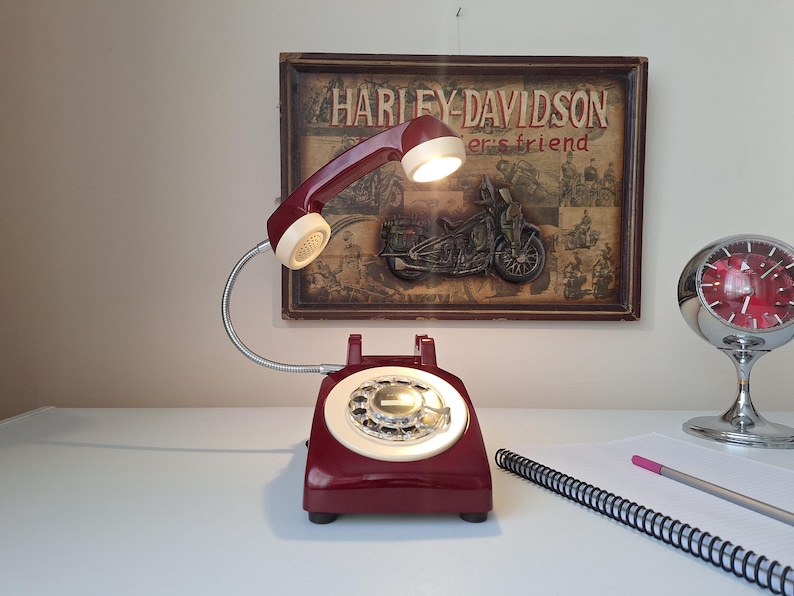 May include: A burgundy rotary phone lamp with a cream-colored dial and handset. The lamp has a flexible silver gooseneck and a small light in the handset. A Harley-Davidson sign is in the background.