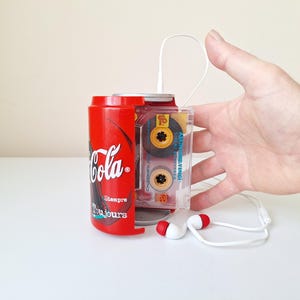 Puede incluir: Una lata roja de Coca-Cola con las palabras "Siempre" y "Toujours" impresas, junto a un reproductor de cintas de cassette transparente. Auriculares blancos con detalles rojos están conectados al reproductor. La lata sostiene el reproductor de cintas.
