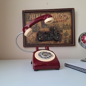 May include: A vintage burgundy rotary phone with a cream-colored receiver and dial. The phone is on a white surface, with a framed Harley Davidson print in the background. A chrome clock and notepad are also visible.