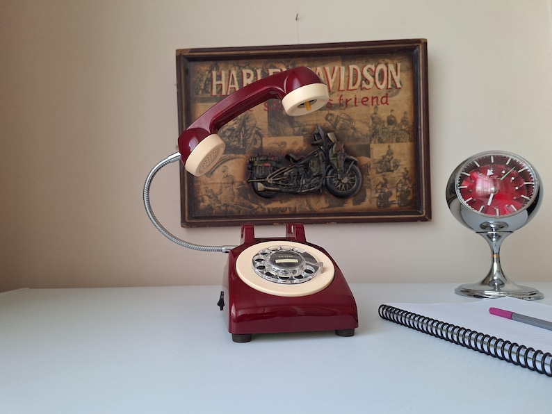 May include: A vintage burgundy rotary phone with a cream-colored receiver and dial. The phone is on a white surface, with a framed Harley Davidson print in the background. A chrome clock and notepad are also visible.