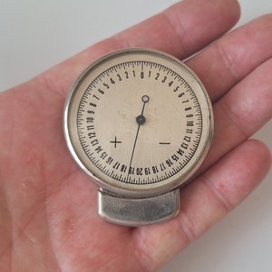 Może przedstawiać: Srebrny, okrągły przyrząd pomiarowy z czarną igłą i białą tarczą. Tarcza ma czarne liczby od 1 do 20, przy czym liczby 15, 16, 17, 18, 19 i 20 pojawiają się dwa razy. Urządzenie ma srebrną metalową obręcz wokół krawędzi.